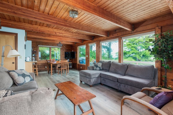Settle into the open-concept living area where warm wood beams, large windows framing lush greenery, and comfortable seating invite you to relax and unwind. Your peaceful retreat awaits.