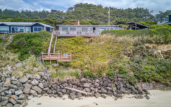Coastal vacation homes nestled among lush greenery with direct beach access via private wooden stairs and deck platform overlooking the sandy shoreline.