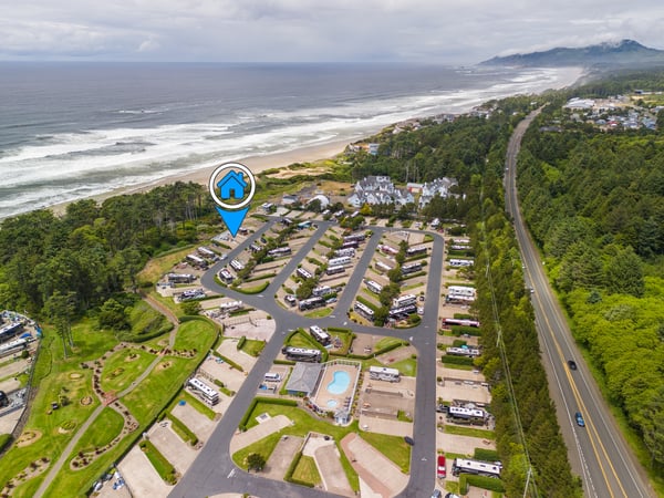 Aerial view of a beachside RV resort nestled between coastal forest and sandy shores, with mountain vistas in the distance. The property features extensive parking and recreational grounds.