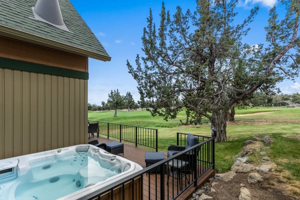 A private deck with hot tub and outdoor seating overlooks a scenic golf course, framed by mature trees and open fairways under blue skies.