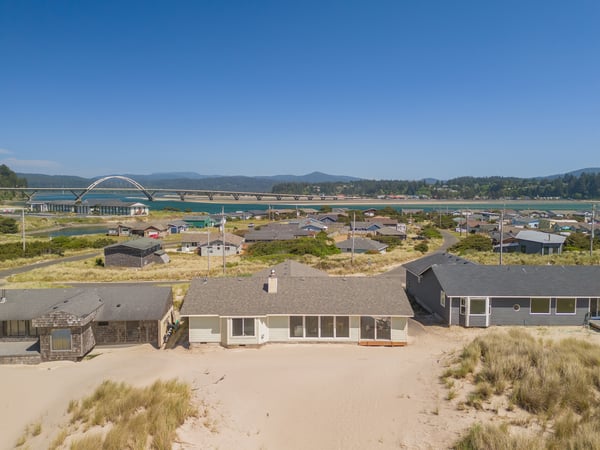 A coastal neighborhood with a distinctive arched bridge spanning turquoise waters, surrounded by forested hills and mountain ranges under clear summer skies.