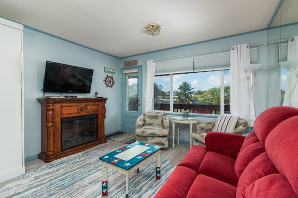 Unwind on the red sofa in your coastal living room, where a fireplace adds cozy warmth and bright windows frame peaceful neighborhood views.