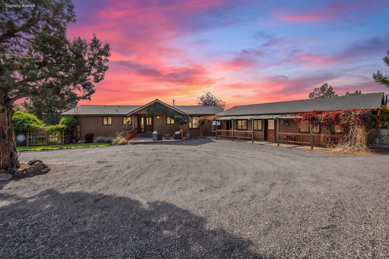 A sprawling ranch-style property sits beneath a stunning sunset, with large mature trees framing the entrance and ample parking space for all your vehicles.