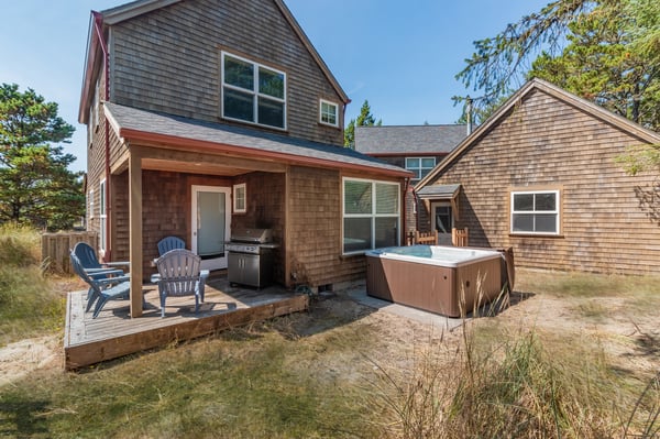 The property features a charming cedar-shingled exterior with a covered patio, outdoor seating, and a private hot tub—perfect for unwinding after a day of exploring the coast.