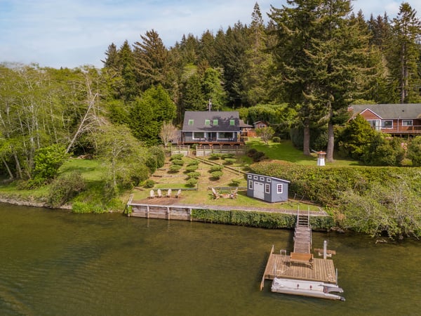 Aerial view of a lakeside property featuring private dock, waterfront lawn, and scenic natural surroundings nestled among tall evergreens and peaceful waters.
