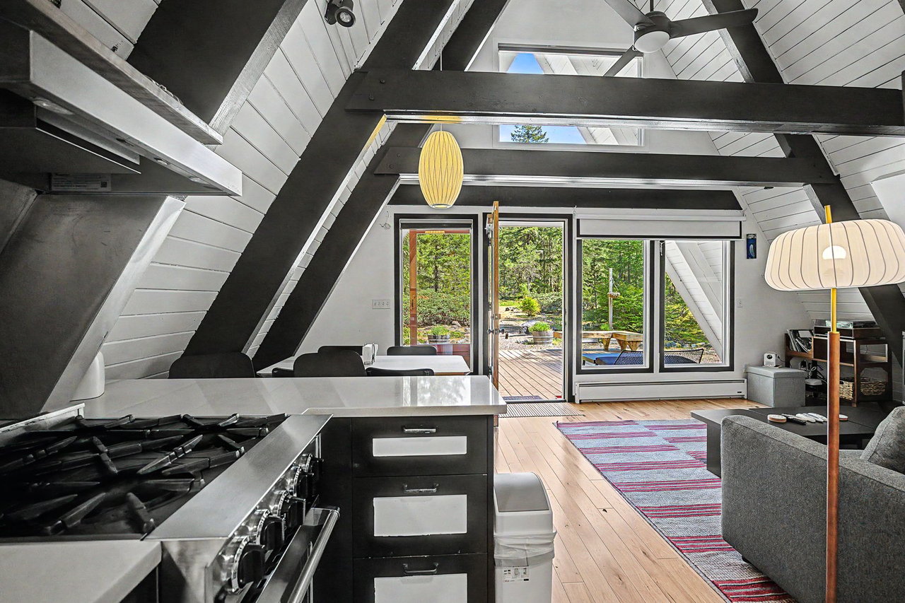 Sunlight streams through skylights onto your open A-frame living space, where modern comforts meet rustic charm. Cook on the sleek gas range while your guests relax on the sofa, all framed by dramatic beams and views of the forest deck beyond.