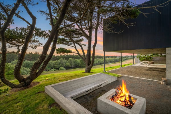 The modern architectural design frames sweeping natural landscapes and a cozy fire pit beneath sheltered living spaces, perfect for peaceful evenings surrounded by majestic trees and open skies.
