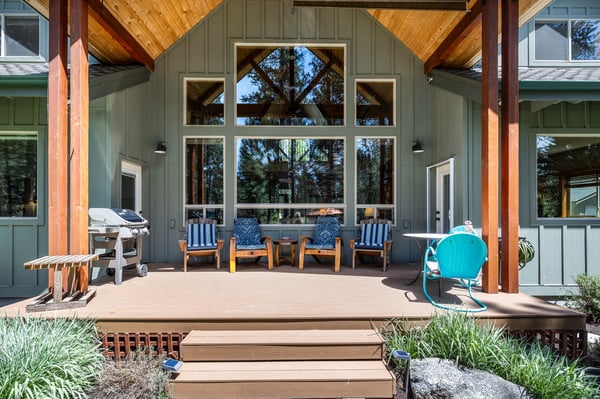 A covered deck surrounded by towering pines, framed by warm wood beams and glass. Gather here with friends around cozy seating or fire up the grill for an outdoor meal.