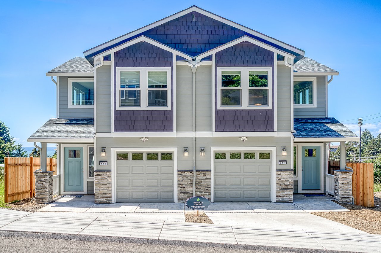 Modern duplex vacation rental featuring spacious two-story design with stone accents, dual garages, and welcoming blue front doors beneath a clear Pacific Northwest sky.