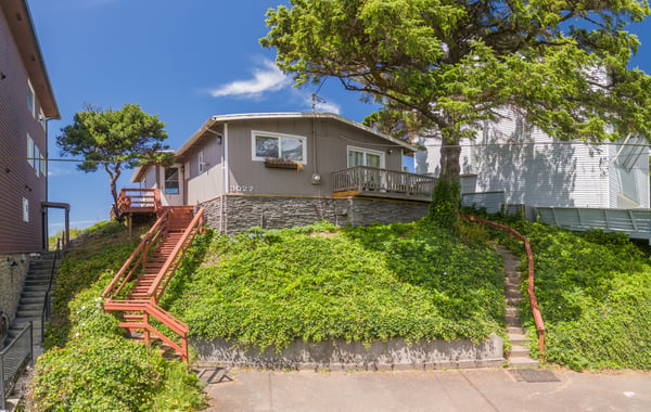 A charming single-story home with a welcoming balcony, nestled on a lush, ivy-covered hillside beneath mature shade trees. Arrive via red railings guiding you up to your peaceful retreat.