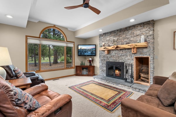 Sink into plush leather sofas before your stacked-stone fireplace, where a ceiling fan whispers overhead and picture windows frame serene woodland views—your cozy retreat awaits.