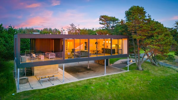 A striking modern architectural retreat set amid lush greenery, featuring two levels of outdoor decks and expansive glass walls that bring nature and comfort together at dusk.