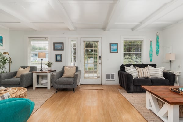 Relax in this spacious living room flooded with natural light through multiple windows. Bright coastal accents and comfortable seating create an airy, inviting atmosphere perfect for unwinding.