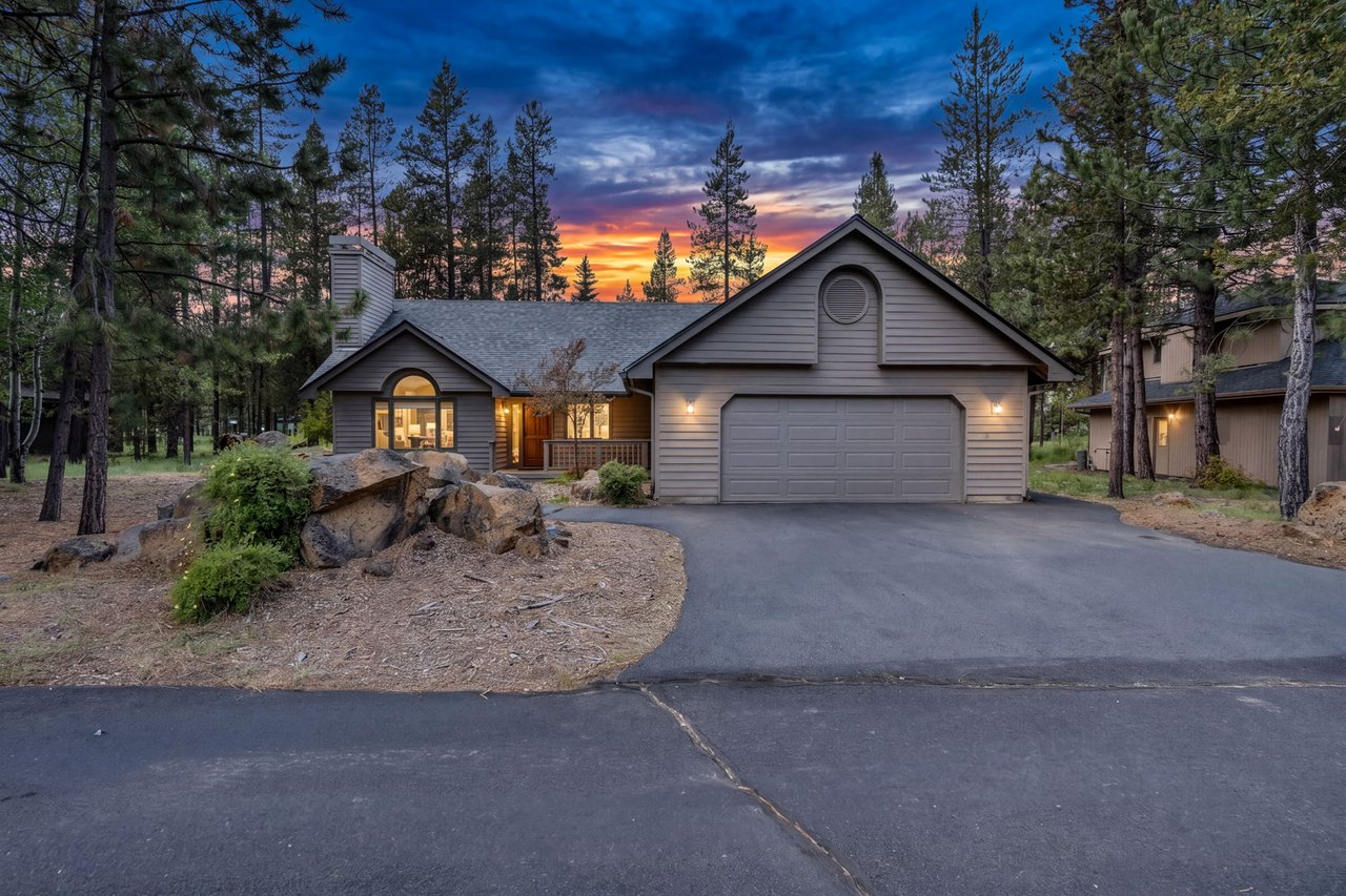 This mountain home nestled among towering pines offers a peaceful retreat with a wide driveway and warm exterior lighting perfect for evening arrivals, all set beneath a stunning twilight sky.