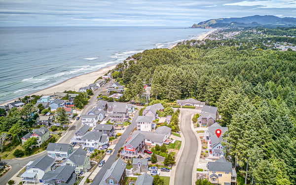 Aerial view of a coastal neighborhood nestled between the ocean and forested hillside, with nearby beach access and mountain backdrop.