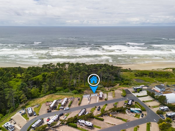 Aerial view of a coastal property nestled between lush forest and sandy beach, with the Pacific Ocean stretching to the horizon. The location marker indicates the property's position within the beachside community.