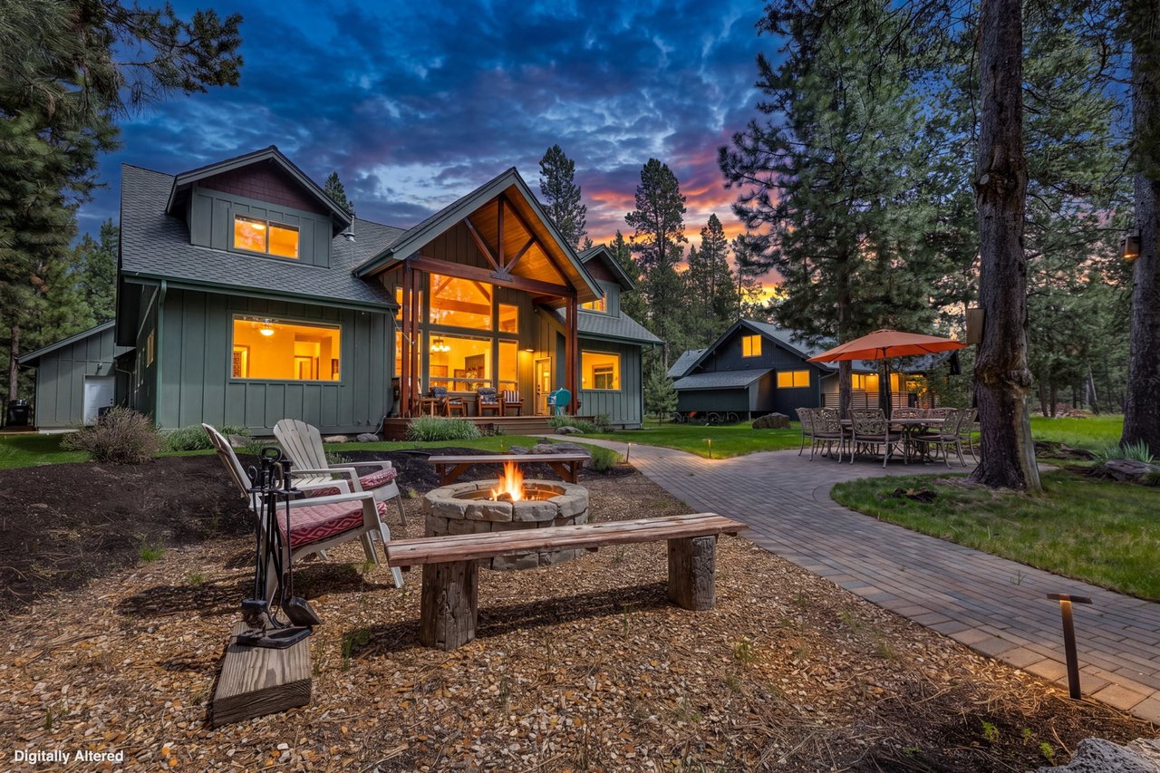 A stunning riverfront retreat surrounded by towering pines, featuring a glowing fire pit and expansive outdoor spaces perfect for evening gatherings under dramatic sunset skies.
