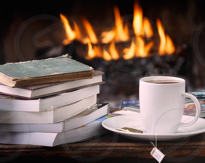 A stack of books next to a cup of a tea in front of a roaring fire